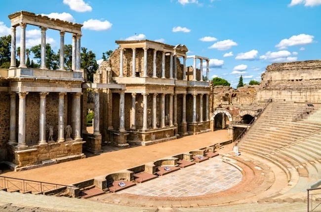 Vista del teatro romano con su imponente scaenae frons de columnas y estatuas, la orchestra semicircular y las gradas de piedra bien conservadas bajo un cielo azul con nubes dispersas. Escenario clásico típico del teatro en Roma.